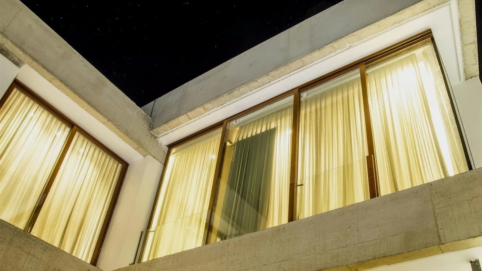 Residencia en Gonnet. Ventanales de gran altura con marco símil madera que se encienden de noche detrás de las cortinas.